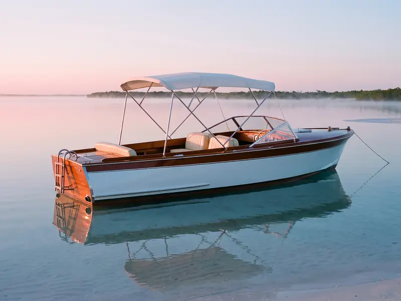 A classic wooden day boat at anchor on a shallow bay at dawn, under a pink sky.