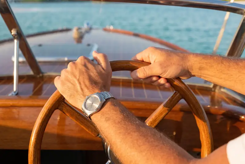 Two tanned hands on a varnished wooden steering wheel, the bay out of focus beyond.
