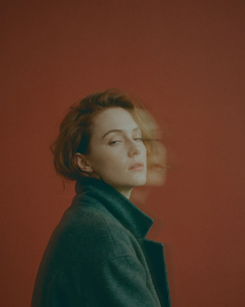 Weight of a Glance: a woman in a charcoal coat looks back over her shoulder against a rust-red wall, a slow-shutter trail beside her face.