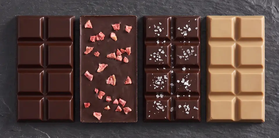 Four chocolate bars side by side on slate: a plain dark bar, a dark bar with pink guava pieces, a dark bar with sea-salt flakes, and a pale café con leche bar