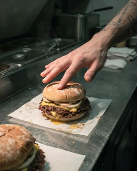 A smash burger on greaseproof paper on a steel counter, a cook's open hand resting on the crown.