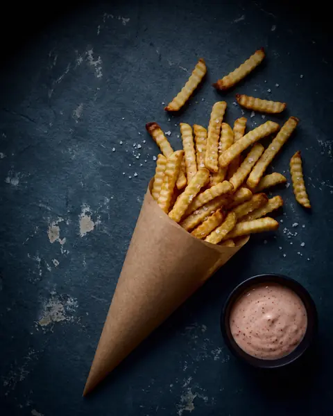 Crinkle-cut fries in a paper cone on dark blue-grey slate, with a small dish of pink sauce.