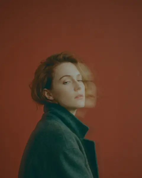 A woman with a short auburn crop in a charcoal coat looks back over her shoulder against a rust-red wall, a smear of motion beside her face.