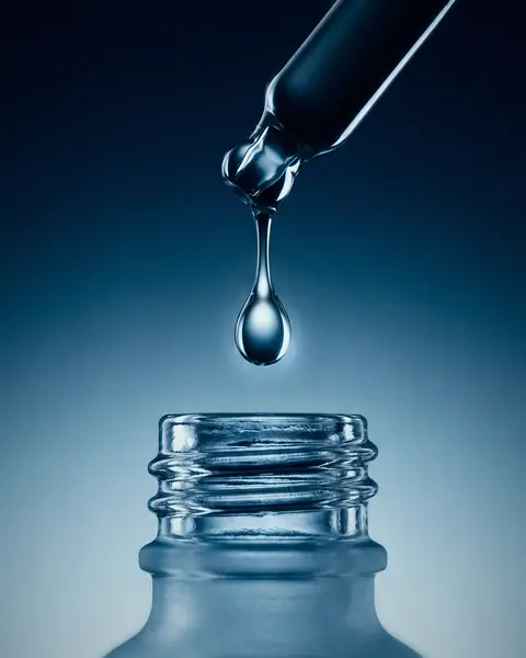 A single drop of clear serum falls from a glass pipette into an open bottle, against cold blue.