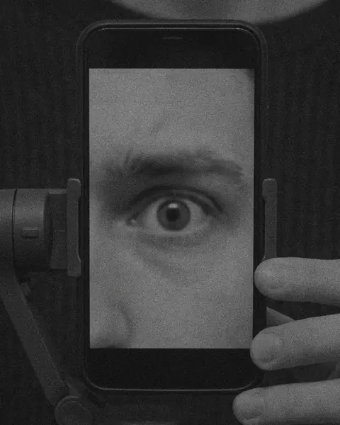 Black and white: two hands hold a phone on a small gimbal, and the screen shows one wide eye.