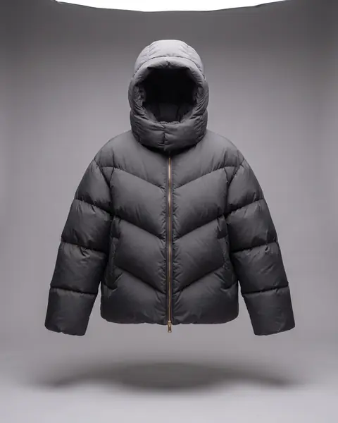 An oversized graphite puffer jacket floats on its own against a mid-grey backdrop: big pillowy baffles, four chevrons meeting at a brass zip, hood up.