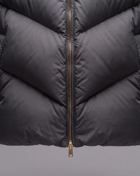Close on the lower front of the jacket: chevron baffles meet at the brass zip, and a flat brass tab hangs at the hem.